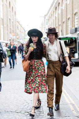 Londra, İngiltere - 17 Temmuz 2019, şık giyimli kadın ve erkek cadde boyunca yürüyor. Adamın beyaz gömleği, bej rengi dar pantolonu ve şapkası var. Kadın çiçekli ve bluzlu uzun bir etek giyiyor..