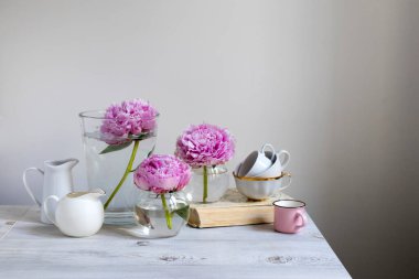 Beyaz bir duvarın arka planında bej ahşap bir masanın üzerinde yuvarlak vazolardaki üç pembe şakayık. Pembe bardak. Metin için boşluk