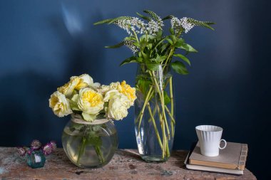 Dar bir vazoda bir buket beyaz Veronica ve yuvarlak vazoda sarı gül, mavi arkaplanda gri bir bankta bir fincan kahve..