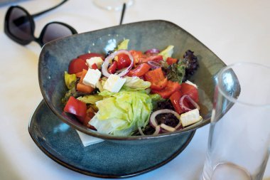 Beyaz bir masada seramik mavi bir kasede Sezar salatası. Güneş gözlükleri yakınlarda.