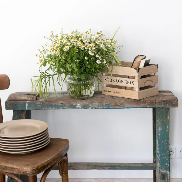 Kitchen decoration. A vase with wild-growing daisies on a shabby bench and a box for storing utensils with books. Wooden Viennese chair with a stack of ceramic plates