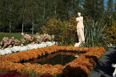 Alçı heykel, sarı ve kırmızı kasımpatı ve ortancalarla kaplı küçük bir gölete yansıyor..