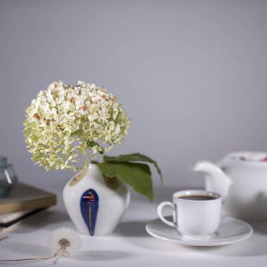Çay içmek için beyaz çaydanlık, fincan ve çay tabağı ve süt testisi. 70 'lerin tarzında küçük bir vazoda kurutulmuş ortanca beyaz masayı süslüyor..
