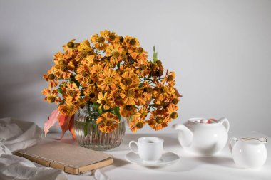 A bouquet of orange helenium with wild grape leaves in a fluted glass vase against a dark blue wall. White teapot, cup and saucer and milk jug for tea drinking. clipart