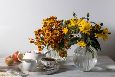 Bir buket turuncu helenyum ve flütlü cam vazoda koyu mavi bir duvara yaslanmış yabani üzüm yaprakları. Çay için beyaz çaydanlık, fincan ve çay tabağı ve süt sürahisi..