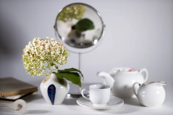 お茶を飲むための白いティーポット カップとソーサーとミルクジュグ 70年代風の小さな陶器の花瓶に緑の干し草が白いテーブルを飾り — ストック写真