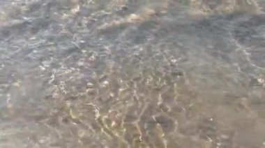 Bir dalga sahile hücum eder ve kumları ve taşları taşıyarak geri döner. Güneş suyun içinde kırılır. Bozulma ve yansıma.