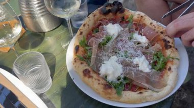 Bir adamın eli, jamon ve mozzarella ile taze yapılmış pizzayı makas kullanarak keser. Masada şarap ve su var..