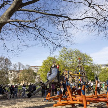 Berlin, Almanya, 5 Mayıs 2025, kalabalık bir şehir oyun alanında güneşli bir gün, büyük, ayrıntılı ahşap ve metal oyun yapısıyla. Çocuklar kaydırak ve ekipman üzerinde tırmanıp oynarken, veliler ve gardiyanlar yakınlarda.