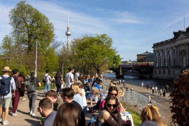 Berlin, Almanya, 5 Mayıs 2025, Berlin 'deki Spree Nehri boyunca kalabalık bir kalabalık güneşli bir günde dinleniyor ve yürüyor. İkonik Fernsehturm (TV Kulesi) uzaktan görülebilir, şehrin üzerinde yükseliyor.