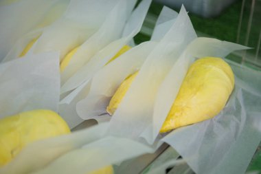 Beyaz kağıda sarılmış taze durian meyveleri, sarı rengini vurgulayan sıcak ışıklarla bir pazarda sergileniyor.