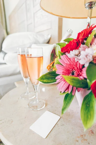 Two glasses of rose champagne and room key card in the upscale hotel room. Dating, romance, honeymoon, valentine, getaway concepts. Vertical. Light warm toning