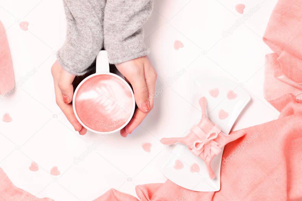 Female hands in grey  knit holding cup of cappuccino. Gift alike dessert with heart shape sprinkles. Flat lay. Valentines concept. Pink textile. Horizontal. Living coral theme - color of the year 2019