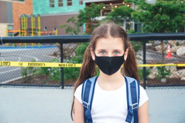 Okul binasının yanında kapalı oyun parkı olan sırt çantalı, bez toz maskeli ilkokul öğrencisi.