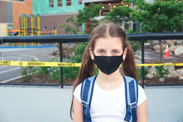 Basisschoolstudent in een stofmasker met rugzak in de buurt van schoolgebouw met gesloten speeltuin Rechtenvrije Stockfoto's