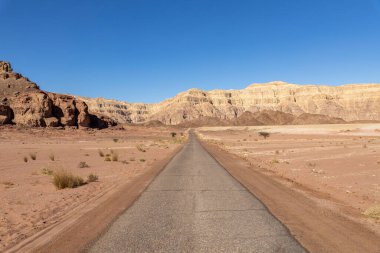 İsrail Çölü Arava 'daki Timna Parkı' ndaki yol manzarası. Mavi gökyüzü, kırmızı-kahverengi dağlar ve taşlar ve birkaç çöl ağacı.