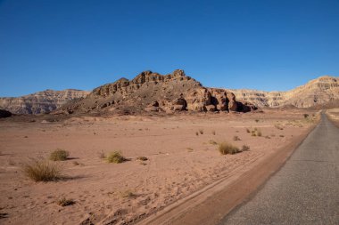 İsrail Çölü Arava 'daki Timna Parkı' ndaki yol manzarası. Mavi gökyüzü, kırmızı-kahverengi dağlar ve taşlar ve birkaç çöl ağacı.