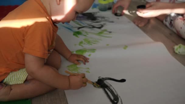 Kind zieht mit schwarzer Farbe und Pinsel auf dem Fußboden sitzend ab. Baby-Scrabble. Handschuss — Stockvideo