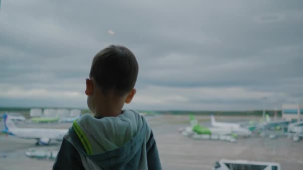 Маленький хлопчик в аеропорту дивиться на машини підтримки і хмарне небо крізь вікно — стокове відео