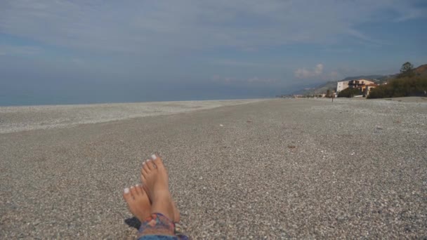 小石のビーチに横たわる女の素足。晴れた日に海のそばを置きます。最初人の眺め — ストック動画