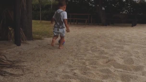 O rapazinho chuta uma bola. Jogando o jogo na praia de areia perto de árvores do parque. Movimento lento — Vídeo de Stock