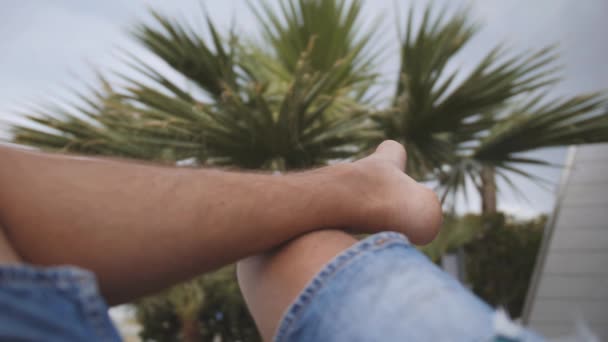 Jambes d'homme pieds nus assis sous un palmier. Un mec détendu en jean. Point de vue — Video