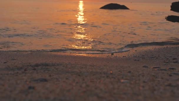 Las olas del mar ruedan en la playa de arena. La puesta de sol brilla en la superficie del agua con amarillo y naranja — Vídeo de stock