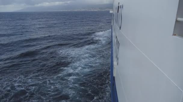 Sida av stora färjan, bow vågor på mörka blå havet och kuststad på horisonten — Stockvideo