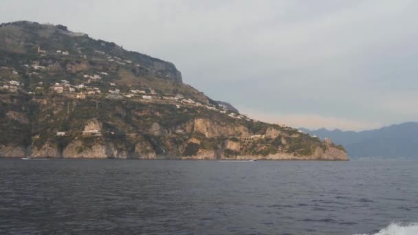 Montaña costera con pueblo en la ladera. Barco a motor flota a lo largo de la costa del mar rocoso, Italia — Vídeo de stock