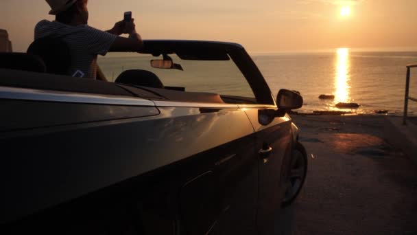 Man takes a photo of sunset seascape by mobile phone from convertible car — Stock Video