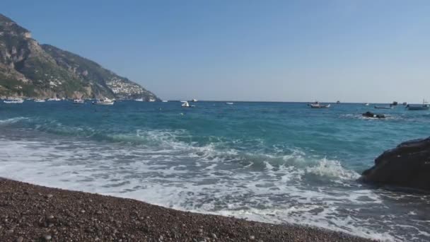 Surf en una playa de guijarros. Las embarcaciones se balancean sobre olas azules cerca de la costa montañosa. Positano, Italia — Vídeos de Stock