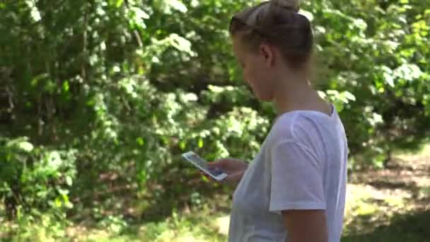 Женщина прокручивает сенсорный экран своего мобильного телефона во время прогулки по парку. Портативный выстрел — стоковое видео