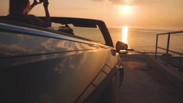 Girl takes a picture of sunset and ocean by smartphone from cabriolet car — Stock Video