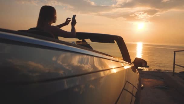 Fille prend une photo de coucher de soleil paysage marin par téléphone mobile de voiture convertible — Video