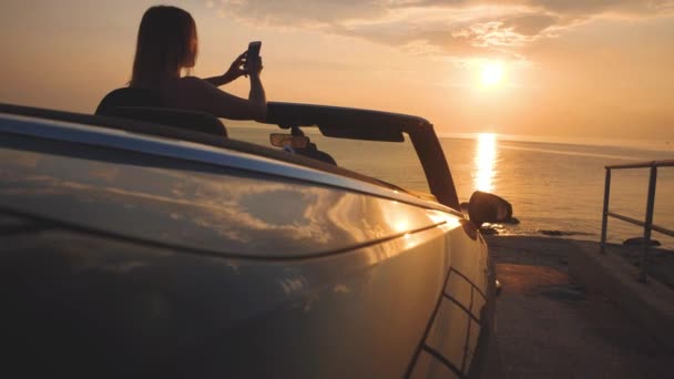 Female driver of luxury car takes s a photo of sunset seascape by cellphone — Stock Video