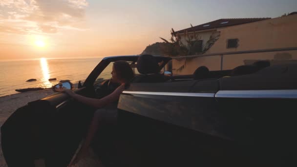 幸せな女性はコンバーチブル車の外に出るし、ビーチに実行されます。海に沈む夕日 — ストック動画