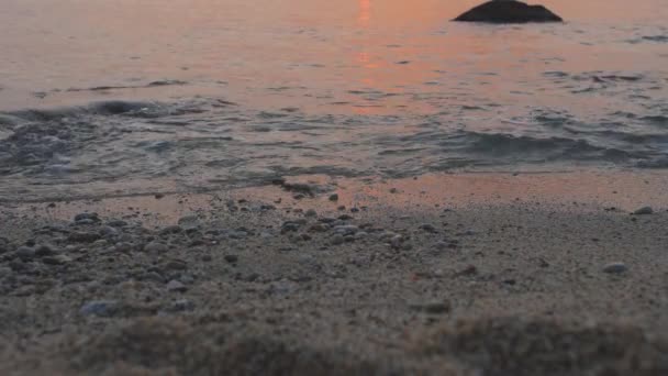 As ondas rolam em areia e pedras de seixo em uma costa do mar. A água reflete a luz do pôr do sol — Vídeo de Stock