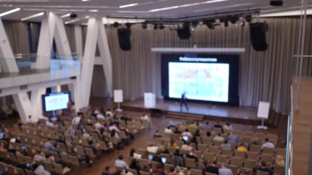 Docent op een podium spreekt voor publiek in collegezaal met bigscreen. Ongericht — Stockvideo