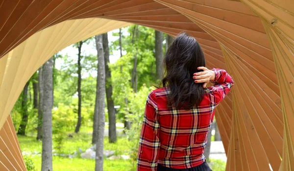 Brunette woman in red shirt standing with her back to the camera and holding her hair in hand inside the wooden tunnel with arches. path to the forest