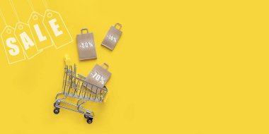 Kağıt torbalı metal alışveriş sepeti, indirimler, sarı arka planda indirim etiketi. Tepeden manzara, düz yerde yatıyordu. Mağazalarda ve internette alışveriş kavramı, satışlar ve Kara Cuma