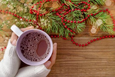 Beyaz kazaklı kadın elleri, yeşil dalları, kırmızı boncukları ve bokehleri olan kahverengi ahşap arka planda, sıcak bir içecekle beyaz bir kupa tutun. Kopya alanı, düz düzen. Mutlu Noeller ve yeni yıl.
