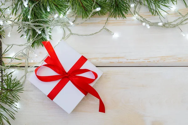 Vánoční přání. Bílá dárková krabice s červenou stuhou na bílém dřevěném podkladu, s borovými větvemi a zářícím věncem. Copyspace, flat lay — Stock fotografie
