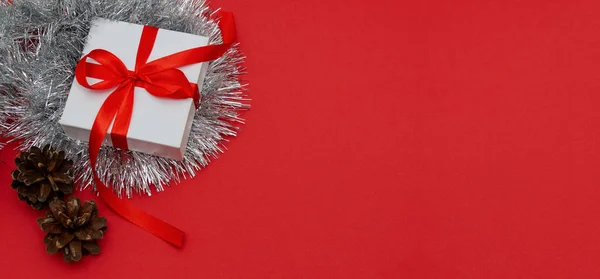 Vánoční prapor. Bílá dárková krabička je svázaná červenou mašlí s větvemi vánočního stromku, borovými kužely a stříbrnou síťovinou na červeném pozadí. Detailní záběr, kopie místa — Stock fotografie
