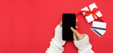 Noel alışverişi. Kadınların elleri beyaz süveter giyer kırmızı arka plandaki akıllı telefondan hediye kutuları ve kredi kartlarının arasında sipariş verir. Pankart, kopyalama alanı, seçici odak