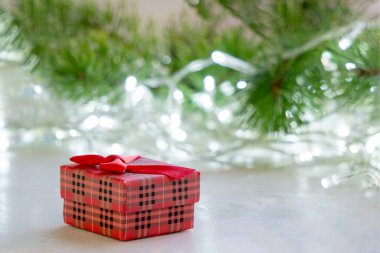 Noel kartı. Masanın ahşap yüzeyindeki kırmızı hediye kutusu köknar dalları ve Noel ışıklarının bulanık arka planına karşı. Mutlu bir yeni yıl konsepti.