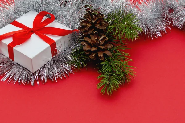 Cartão de Natal. Caixa de presente branco amarrado com um arco vermelho com galhos de árvore de Natal, cones de pinho e malha de prata em um fundo vermelho — Fotografia de Stock