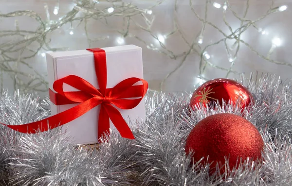 Cartão de Natal. A caixa de presente branca é amarrada com um arco vermelho com bolas de Natal vermelhas em um fundo branco com uma guirlanda espumante e ouropel de prata — Fotografia de Stock