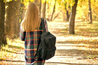 Sırtında kitaplar için siyah bir sırt çantası olan sokakta genç bir kız parkta bir patika boyunca yürüyor. Okul, enstitü ve karantinadan sonra çalışma kavramı..