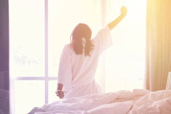 Woman stretching hands on bed after waking up while sitting on bed, entering a day happy and relaxed after good night sleep
