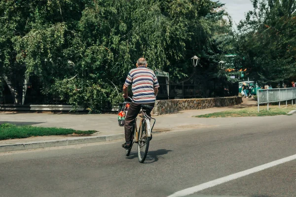 An elderly man with a receding hairline in a striped t-shirt rides his bike off the road to a park in daylight in a populated project